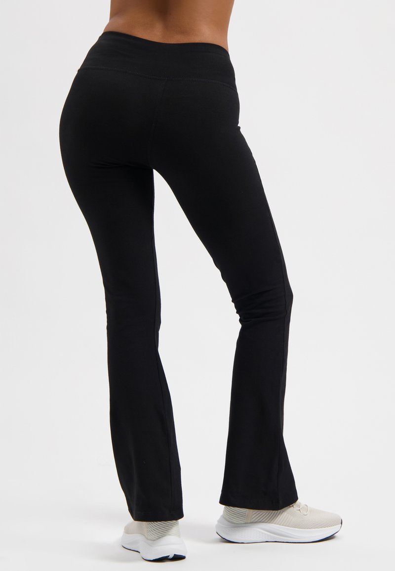 Schwarze, dehnbare Schlaghosen mit hohem Bund, weichem Material und glatten Nähten, getragen mit hellen Sportschuhen.