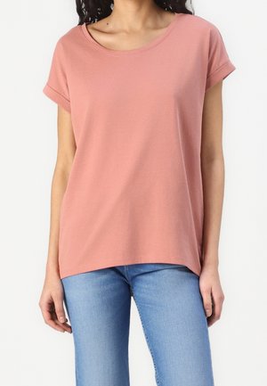Vrouw draagt een effen stoffig roze korte mouw T-shirt en lichtblauwe spijkerbroek, staand tegen een witte achtergrond.