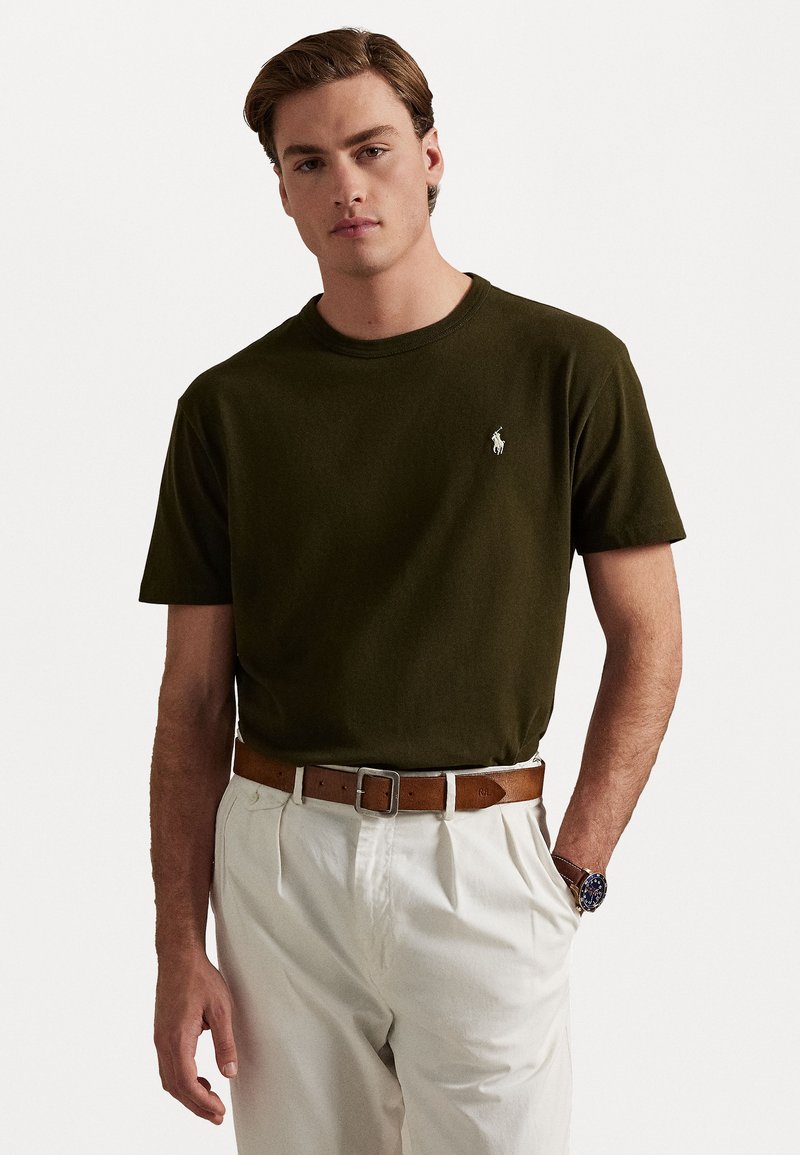 Mörkgrön bomull t-shirt med rundad halsringning och korta ärmar, prydd med en liten vit broderad logotyp på vänster bröst. Bärs med vita byxor.