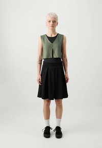 Zelena pohodniška jakna preko črne majice, v kombinaciji s črno plisirno krilo. Model nosi bele nogavice in črne čevlje. Preprosta zasnova in čiste linije.