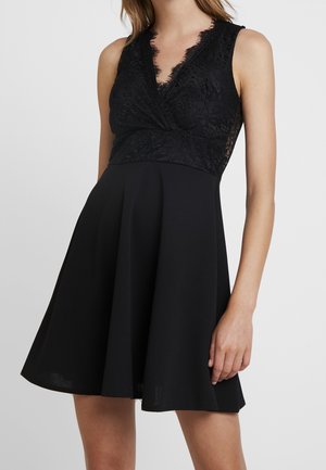 Czarna sukienka bez rękawów z koronkowym dekoltem w kształcie litery V i rozkloszowanym dołem, noszona przez osobę, widziana od ramion do połowy uda na jednolitym tle.