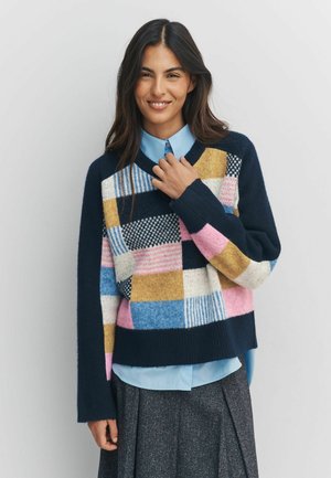 Kvinna som ler, iklädd en flerfärgad lappstickad tröja över en ljusblå skjorta med krage och en mörkgrå plisserad kjol, stående mot en enfärgad bakgrund.