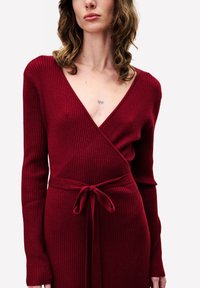 Rode geribbelde wikkeljurk met lange mouwen, diepe V-hals en een stoffen riem die in de taille is gestrikt. Getextureerd materiaal geeft diepte aan het ontwerp.