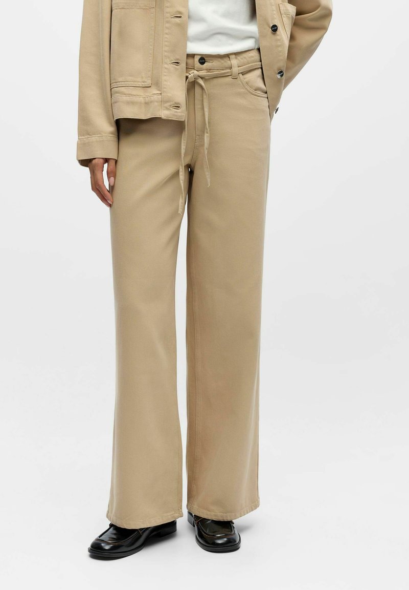 Bēšas krāsas platām kājām bikses no mīksta auduma, ar auklu regulējamu jostasvietu, diskrētām sānu kabatām un gludu, vienkāršu virsmu.