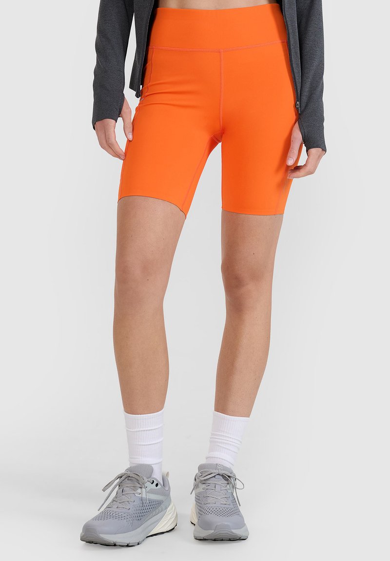 Personne portant un short moulant orange vif, une veste zippée grise, des chaussettes blanches montantes et des chaussures de sport grises, debout sur un fond neutre.