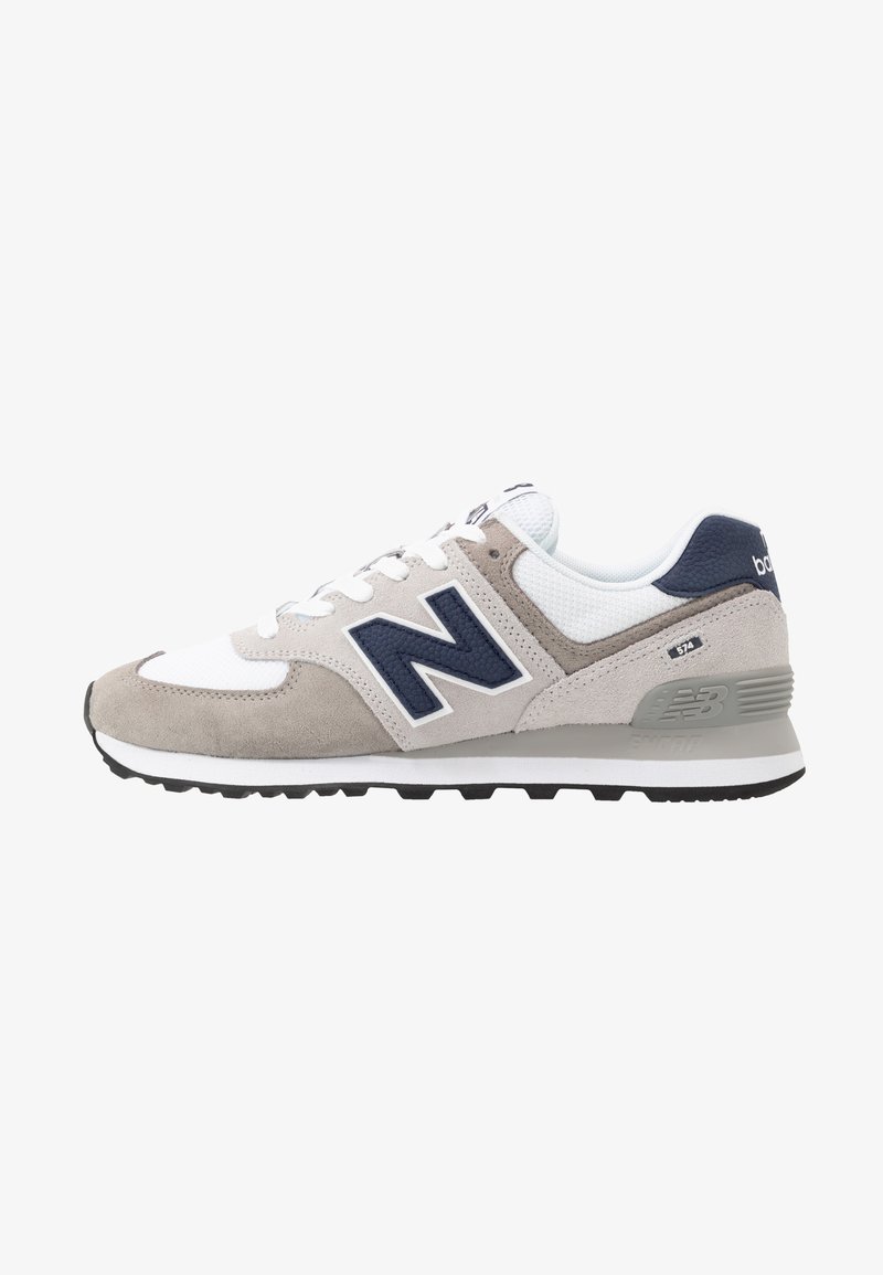 Baskets basses New Balance 574 blanches et grises avec logo bleu marine, tige en daim et mesh, et semelle rembourrée avec semelle extérieure noire.