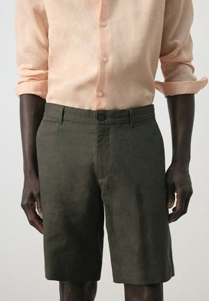 Uomo che indossa una camicia pesca a maniche lunghe arrotolate e pantaloncini verde oliva scuro fino al ginocchio, in piedi davanti a uno sfondo chiaro e semplice.