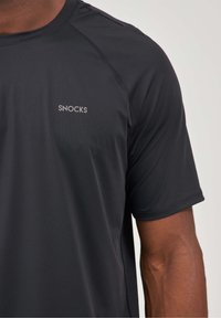 T-shirt de sport noir en matériau léger, avec des manches courtes et un petit logo "SNOCKS" sur la poitrine.