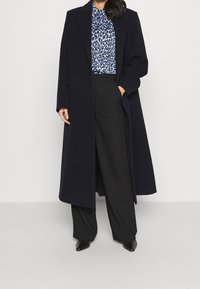 Long manteau en laine mélangée bleu marine à double boutonnage, porté par-dessus un chemisier à imprimé léopard bleu et un pantalon large noir. Chaussures noires à talons.