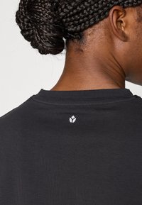 Μαύρο t-shirt με στρογγυλή λαιμόκοψη και κοντά μανίκια, με μικρό λευκό λογότυπο στην πλάτη κοντά στον λαιμό. Ομαλή υφή υφάσματος.