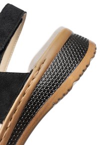 Schwarze Wildledersandale mit braun-schwarzer, strukturierter Gummisohle, sichtbaren Nähten und gepolstertem Fußbett auf weißem Hintergrund.