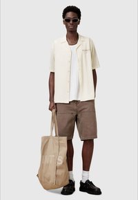 Conjunto casual com uma camisa de manga curta crème, calções cargo castanhos, sapatos pretos, meias brancas e uma bolsa tote bege com um logo.