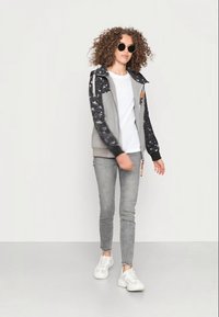 Casaco cinzento com fecho éclair e mangas pretas cintilantes, combinado com uma t-shirt branca e jeans skinny cinzentos, finalizado com sapatilhas brancas.