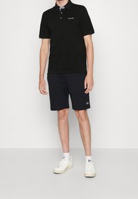 Polo shirt nera a maniche corte con colletto, abbinata a pantaloni corti blu navy. Sneakers bianche completano l'outfit, con un piccolo logo sui pantaloni.