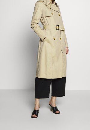 Gabardina beige con cinturón y botones dorados, combinada con pantalones negros de pierna ancha y tobillo descubierto, y mules negros con tacón y punta abierta.