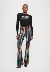 Čierny dlhý rukáv s logom "Moschino Jeans"; farebné, vzorované rozšírené nohavice s zipsami na čiernom pozadí; vysoké podpätky.