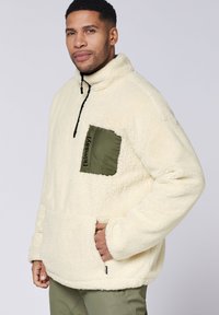 Mann in cremefarbenem Fleece-Pullover mit olivgrünem Brusttasche und Kragenreißverschluss, der mit einer Hand in der Tasche vor einem hellen Hintergrund steht.