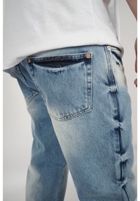 Los jeans de mezclilla azul claro deslavado cuentan con un diseño de cinco bolsillos, remaches de cobre y un sutil desgaste en la parte posterior, con un corte relajado.