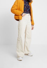 Cremekleurige wijde corduroy broek gecombineerd met een lange mouwen top met bloemenprint en een feloranje donsjack. Witte sneakers maken de outfit compleet.