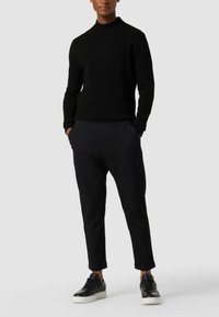 Schwarzer strukturierter Strickpullover mit hohem Kragen, kombiniert mit maßgeschneiderten schwarzen Hosen und schwarzen Sneakers mit weißen Sohlen.