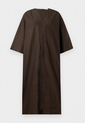 Femme portant une robe midi ample marron foncé à manches larges, tenant une petite pochette tissée et portant des chaussures plates noires.