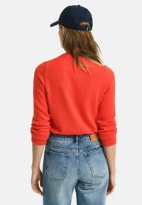 Red knitted sweater with ribbed cuffs, worn over blue high-waisted denim jeans. Navy cap completes the casual outfit.