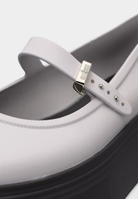 Λευκό παπούτσι Mary Jane με ρυθμιζόμενο λουράκι και χρυσή αγκράφα με χαραγμένο το όνομα "Melissa" πάνω από παχύ μαύρο πέλμα.