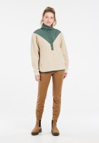 Fleece pullover in beige met een groene schouderstuk, met een hoge kraag, een half-knopsluiting en voorkeurszakken. Bruine broek en enkellaarsjes.