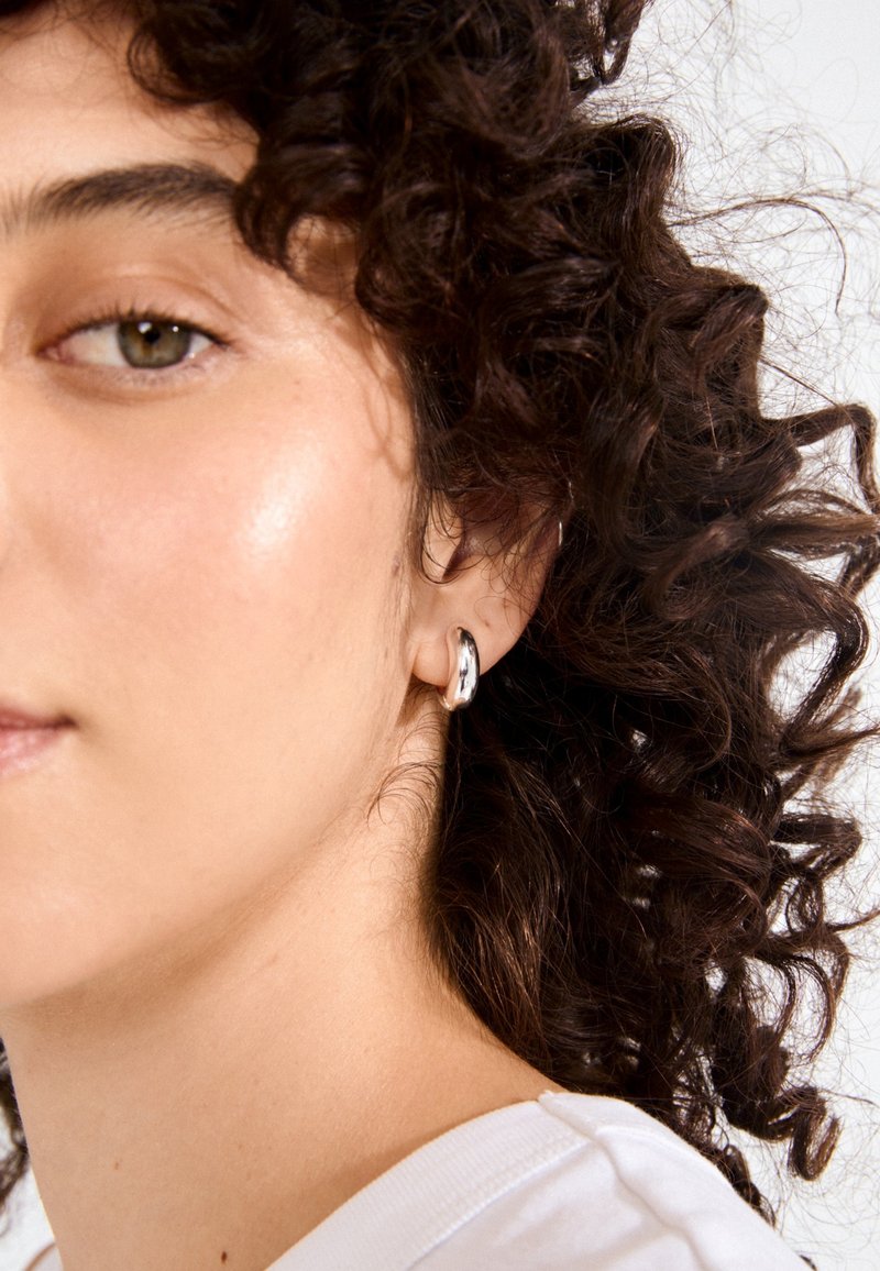 Sidabrinė apyrankė su lygiu, atspindinčiu paviršiumi. Kreipimo dizainas puikiai dera prie garbanotų plaukų. Fonas yra paprastas ir neutralus.