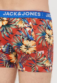 Red floral patterned boxer shorts with a blue elastic waistband featuring "JACK & JONES" logo, made from stretchy fabric for comfort.