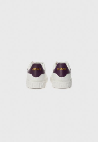 Adidași albi cu toc violet, având un logo "Versace" aurit. Talpă texturată cu un model geometric. Design și formă modernă.