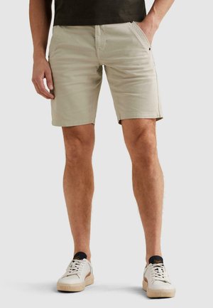 Mand iført beige afslappede shorts og hvide sneakers, stående med den ene hånd i lommen mod en ensfarvet baggrund.