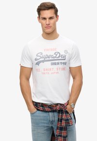 Camiseta blanca de algodón con estampado gráfico en gris y coral. Llevada con pantalones vaqueros azules, una camisa de cuadros atada a la cintura y un reloj dorado.