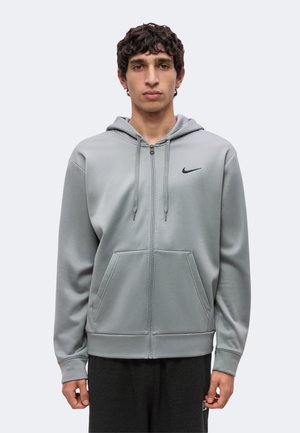 Mladý muž stojaci, oblečený v svetlosivej mikine Nike na zips s kapucňou a prednými vreckami, v kombinácii s tmavými nohavicami na jednofarebnom pozadí.