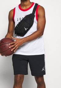 Vit mesh linne med röd kant, svarta shorts och en svart axelväska med vit Jordan-logotyp. Håller en brun basketboll.