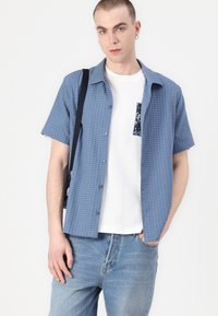 Short-sleeve, textured blue shirt with a collar, button closure, and a patterned pocket, worn over a white t-shirt and denim jeans.