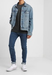 Veste en denim bleu clair portée sur un pull gris foncé, associée à un jean bleu ajusté et des baskets montantes blanches avec les revers retroussés.