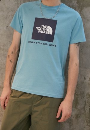 Lichtblauw t-shirt met korte mouwen en een vierkant logo in marineblauw, voorzien van de tekst "NOOIT STOPPEN MET VERKENNEN" eronder. Zachte stof, relaxte pasvorm.