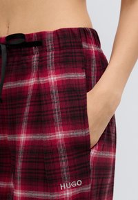 Κόκκινο και μαύρο καρό παντελόνι πυτζάμας με κορδόνι στη μέση, πλαϊνή τσέπη και λευκό λογότυπο HUGO στο αριστερό πόδι. Μαλακή υφή.