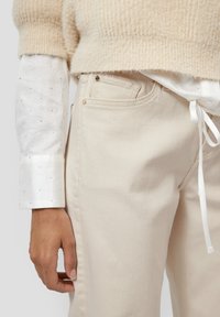 Kremowe, wysokiej talii dżinsowe spodnie o gładkiej fakturze i bocznych kieszeniach. Połączone z beżowym swetrem z dzianiny oraz przejrzystą białą bluzką.