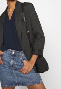 Donna che indossa un blazer grigio a righe sottili, una blusa blu navy, una gonna di jeans e una borsa a tracolla in pelle nera, con le mani nelle tasche, tagliata alle spalle e alle cosce.