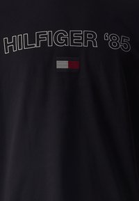Camiseta de algodón negra con "HILFIGER '85" en texto blanco y un pequeño parche con logo en franjas rojas, blancas y azules.