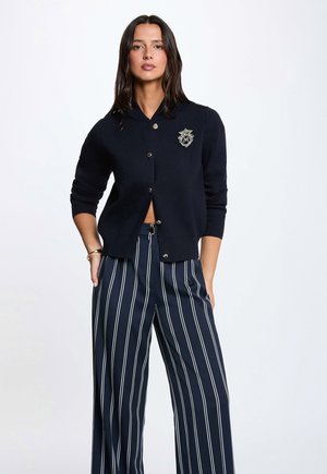 Vrouw draagt een zwarte buttoncardigan met embleem en marineblauwe wijde broek met witte verticale strepen, staand met handen in de zakken.