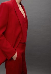 Costume rouge comprenant un blazer ajusté avec des épaulettes et un gilet cintré à boutons, associé à un pantalon slim. Texture lisse.