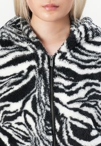 Juodai baltas zebra rašto zip-up fliso džemperis su minkšta tekstūra, su aukštu apykakle ir matomu užtrauktuku.
