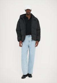 Must must jacket kapuutsiga, millel on lukku, kantud musta polo särgi kohal. Koos helesiniste laiade püksidega ja mustade jalatsitega.