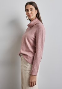 Ljusrosa polotröja med ribbad halsringning och manschetter, kombinerad med ljusa kordflöjlsbyxor, sedd från sidan mot en neutral bakgrund.