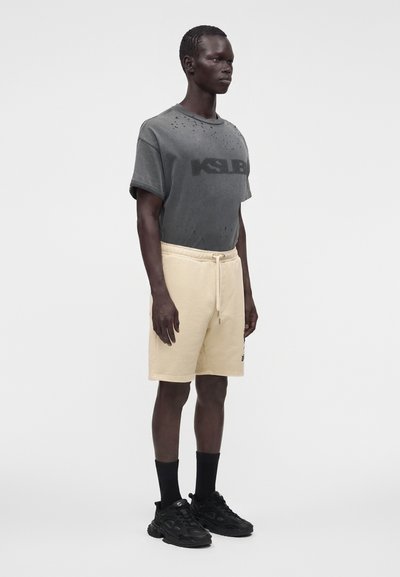 Homme debout portant un t-shirt gris foncé usé, un short beige avec cordon, des chaussettes noires et des baskets noires sur un fond uni.