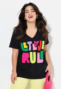 Sort V-hals T-shirt med flerfarvede blokbogstaver, der står "LAD KÆRLIGHEDEN REGERE." Model iført gule bukser og holder en lyserød taske.