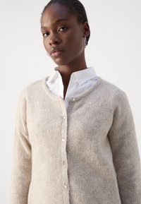 Gilet beige clair avec une texture douce et côtelée. Il possède un col rond et des boutons en perles sur le devant. Superposé sur une chemise blanche.
